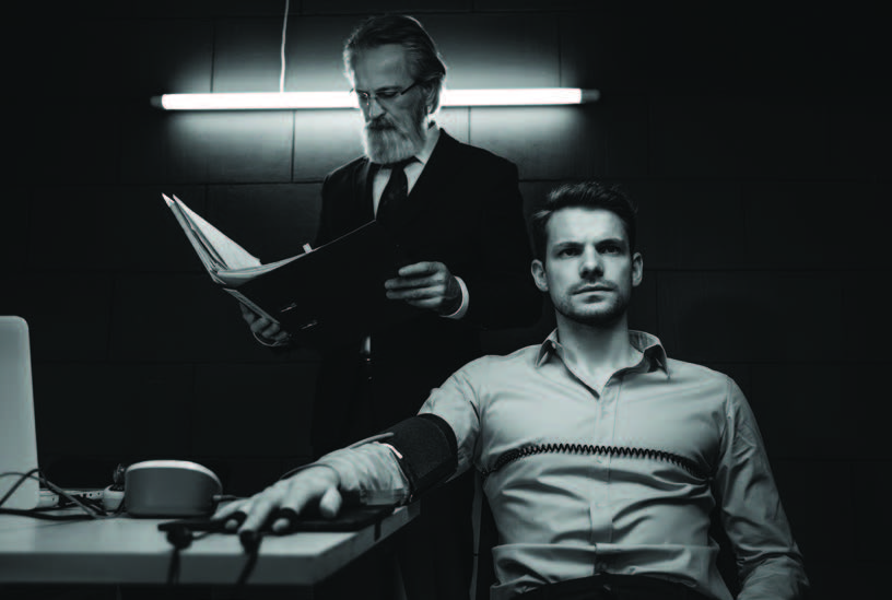 Photograph of one man looking as if he is being interrogated while another man reads from a binder behind him