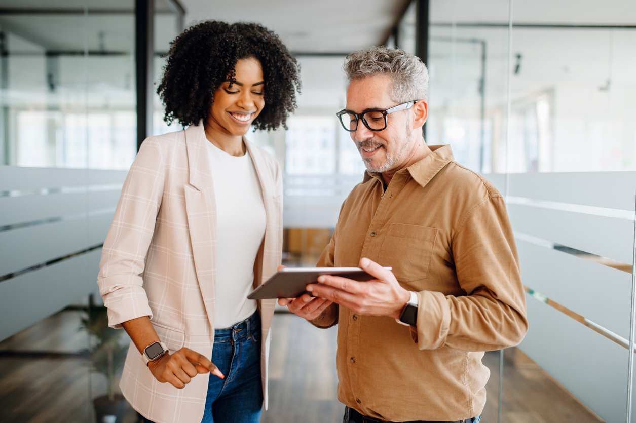 Stock photo of two cheerful colleagues, representing independent agents, share a tablet screen, collaborating on options from multiple companies to help clients make an informed decision.